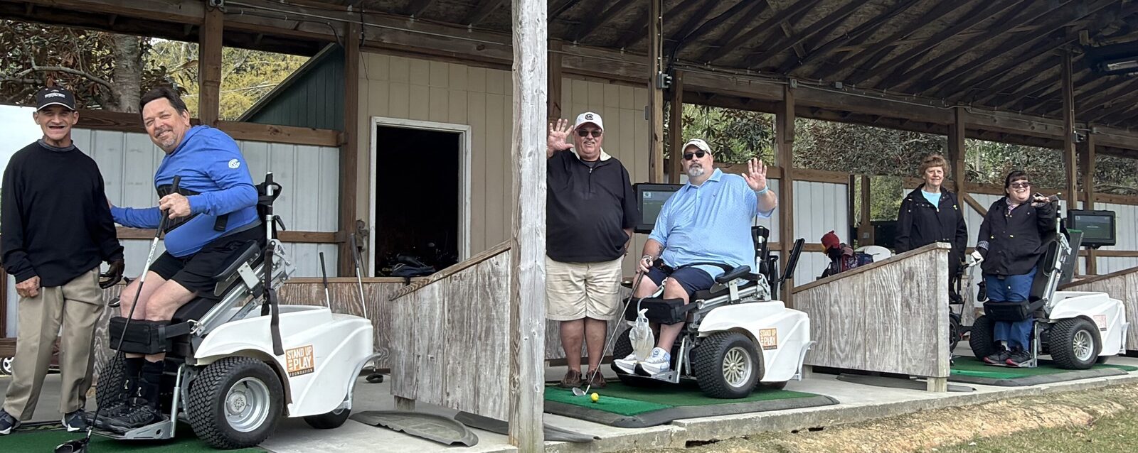 Participants using Stand Up and Play adaptive golf carts at an MTTG community clinic, waving and smiling alongside Marty, an MTTG assistant instructor, and a parent/care provider under the driving range shelter