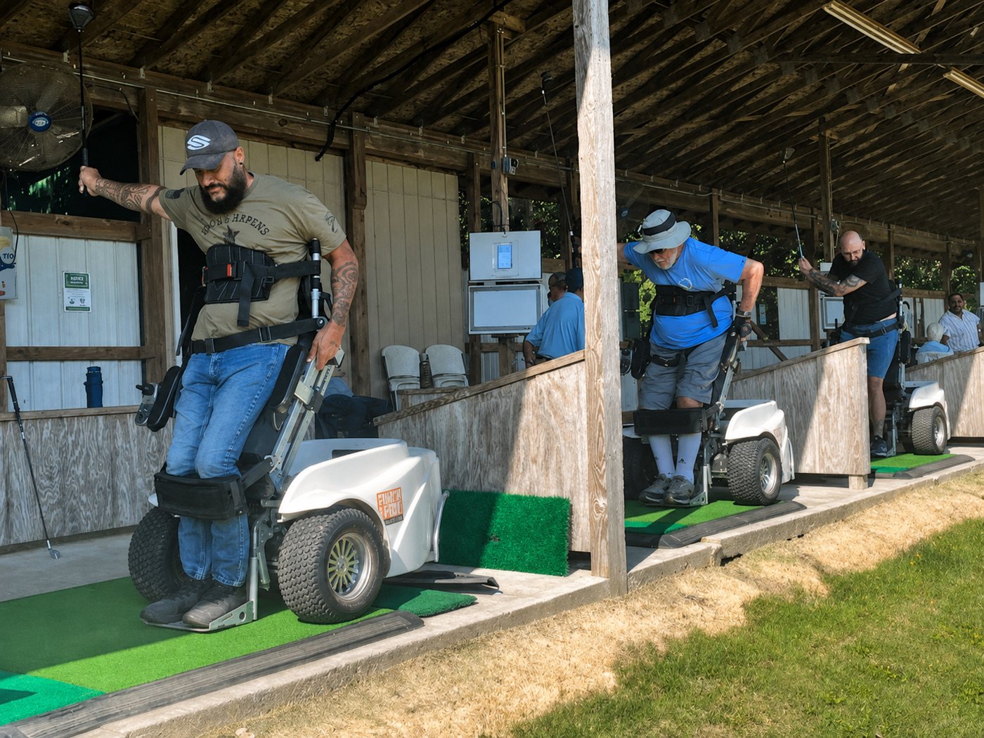 Participants using adaptive standing mobility devices to hit golf balls under a covered driving range